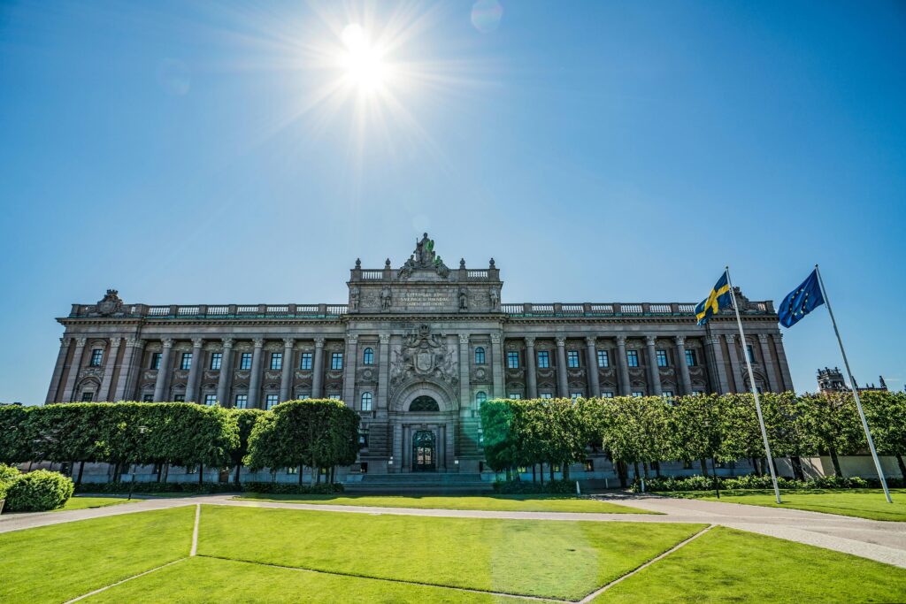 Front view of the Riksdag building with Swedish and EU flags on a sunny day.