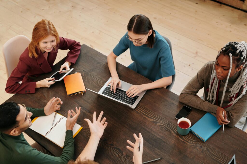 High-angle view of diverse team discussing and working on laptops and notepads around wooden table.