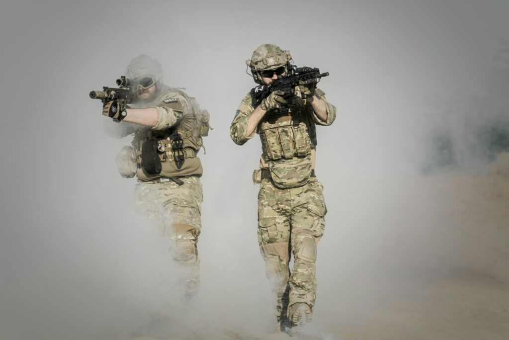 Two soldiers in camo uniforms with weapons advancing through a smoke-covered field, showcasing military action.